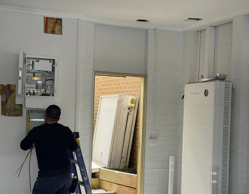 An electrician in black shirt working on electrical panel installation next to white home battery energy storage with solar system in residential garage