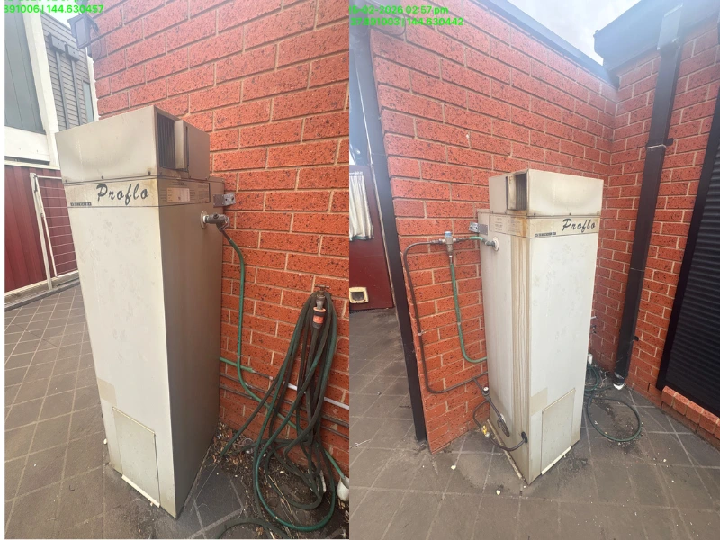 Old Proflo hot water system installed against a red brick wall outdoors, with a green garden hose coiled nearby